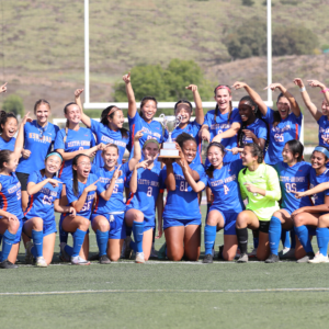 The Pomona-Pitzer women's soccer team gather with their coaches on the 场 in two rows while raising their hands triumphantly. 中间的一名球员举着奖杯. 球队身穿蓝色制服，配以白色和橙色.